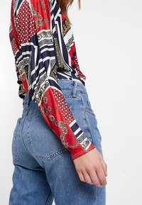 Rød og marineblå mønstret langærmet top med geometriske designs og kædemotiver, parret med højtaljede blå denim jeans.