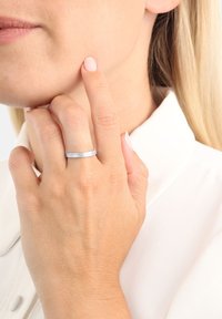 Anello d'argento con superficie texturizzata, indossato su una mano che tocca il viso. Sullo sfondo si intravede una camicia bianca semplice. Il tono della pelle appare naturale.