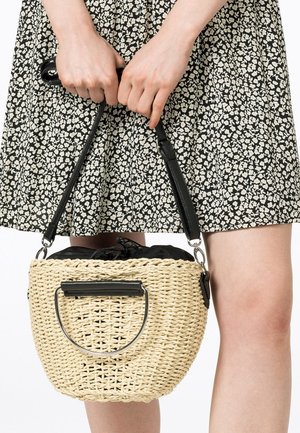 Mains tenant un sac à main en cuir tressé beige avec des poignées en cuir noir, porté avec une jupe fleurie noire et blanche.