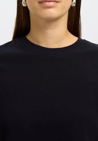 Nahaufnahme einer Person, die ein schlichtes schwarzes T-Shirt mit Rundhalsausschnitt trägt, mit langen braunen Haaren und kleinen silbernen Creolen.