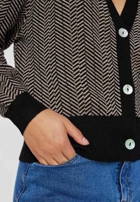 Pletený kardigan má černo-béžový rybí kost vzor, výstřih do V a knoflíky připomínající perly. Manžety a lem jsou černé.