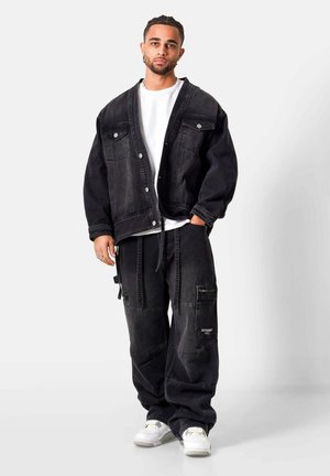 Junger Mann mit geflochtenem Haar, trägt eine übergroße schwarze Jeansjacke, passende Cargo-Hosen, ein weißes T-Shirt und weiße Sneakers, steht vor weißem Hintergrund.