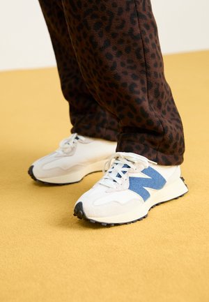Hvide sportssko med blå accenter og teksturerede paneler, kombineret med mørkebrune leopardmønstrede bukser, på en gul baggrund.