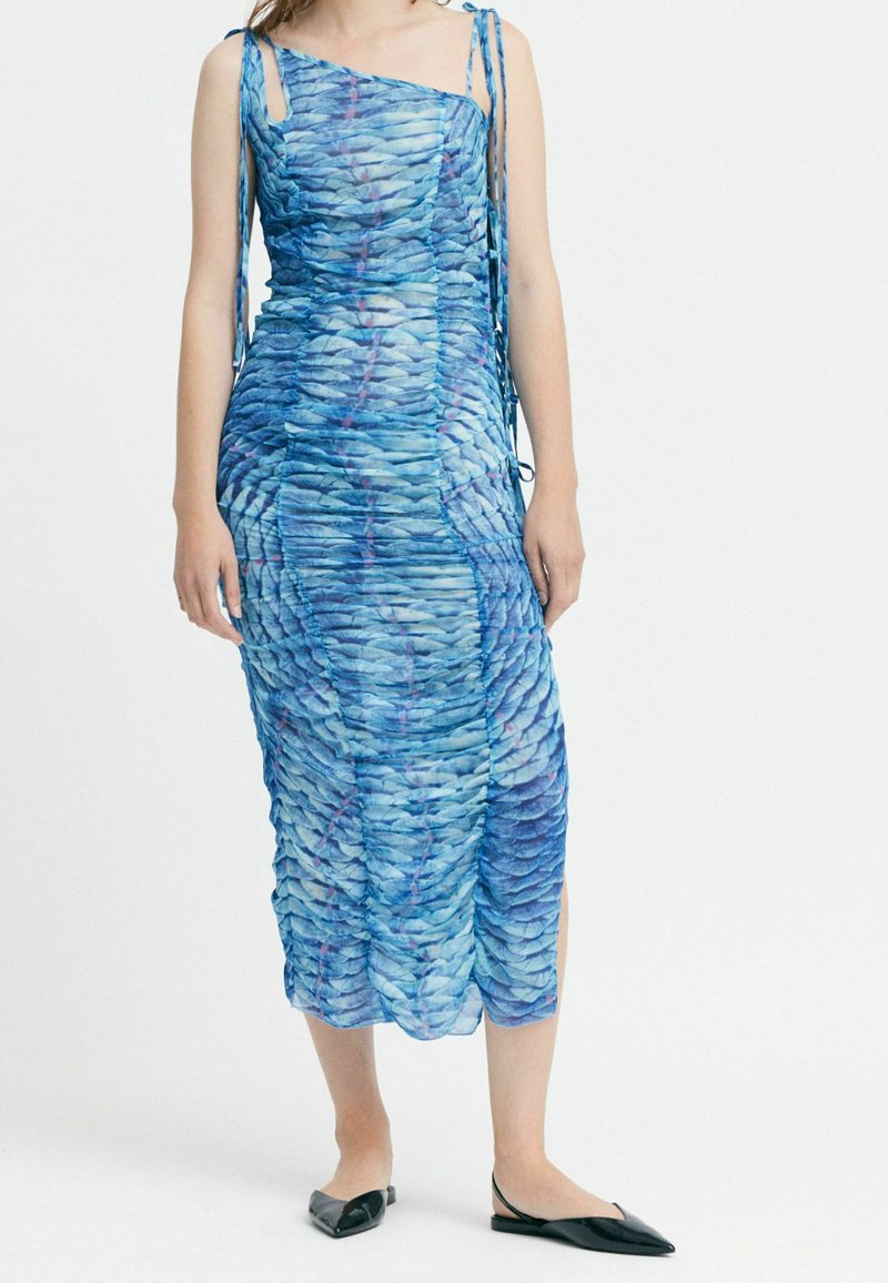 Blaues Midi-Kleid aus strukturiertem, gerafftem Stoff mit wellenförmigen Mustern. Ein Schulterträger und seitliche Schlitzchen. Schwarze flache Schuhe.