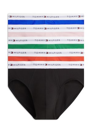Ung mand står barfodet iført sorte Tommy Hilfiger briefs med hvid taljebånd og kigger mod venstre mod en hvid baggrund.