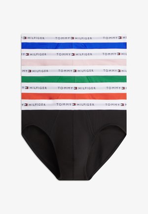 Jeune homme debout pieds nus portant un slip noir Tommy Hilfiger avec une ceinture blanche, regardant vers sa gauche sur un fond blanc.