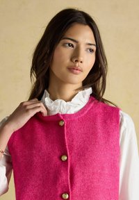 Leuchtend pinke Wollweste mit rundem Ausschnitt und vier goldenen Knöpfen. Strukturierter Stoff am Körper; darunter eine weiße, mit Rüschen versehene Bluse.