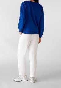 Persona con un sudadera azul, pantalones blancos y zapatillas blancas, de pie con las manos en los bolsillos contra un fondo claro y sencillo.