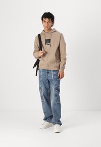 Beige hoodie med en brystlomme, parret med blå denim cargo pants, der har flere lapper og hvide accenter. Hvide sneakers fuldender looket.