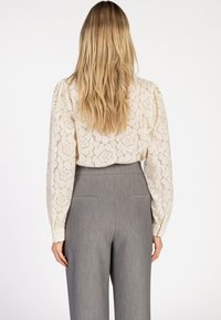 Vrouw met lang blond haar, gekleed in een crèmewitte bloemenkant blouse en hoge, grijs op maat gemaakte broek, met haar rug naar de camera gekeerd.
