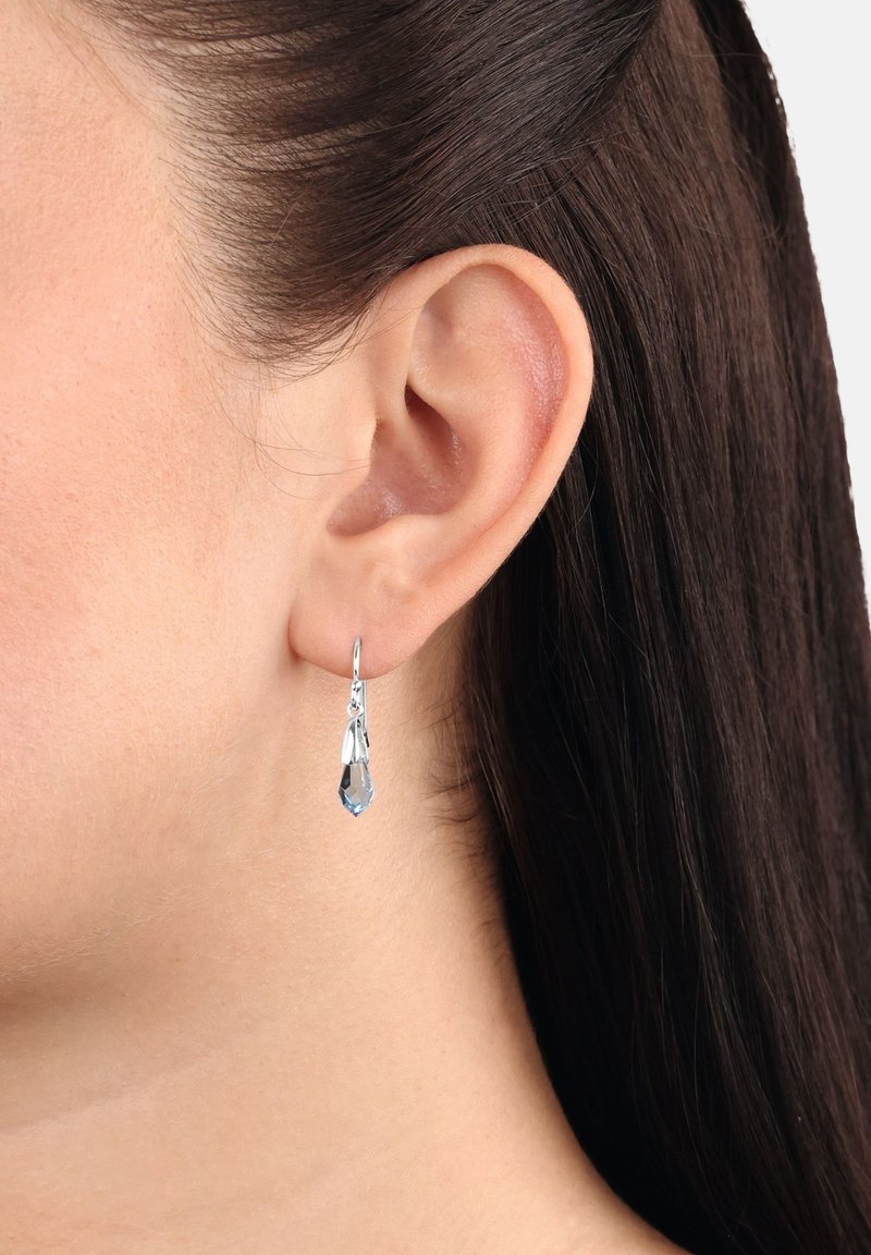 Oreille gauche avec des cheveux bruns foncés et lisses tirés derrière, porte une boucle d'oreille argentée en crochet avec un pendentif en cristal clair en forme de goutte.