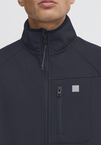 Sötétkék softshell dzseki magas gallérral, cipzáras elülső résszel és oldalsó zsebbel; a mellkason egy kis csíkos logó látható. Sima felület.