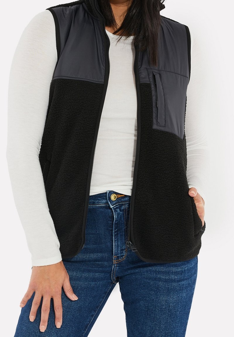 Femme portant un gilet noir sans manches avec une fermeture éclair à l'avant et une poche poitrine sur une chemise blanche à manches longues et un jean bleu.
