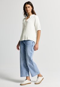 Blouse blanche texturée avec un col en V et des volants, associée à un jean bleu clair à jambes larges et des baskets blanches avec des accents noirs.