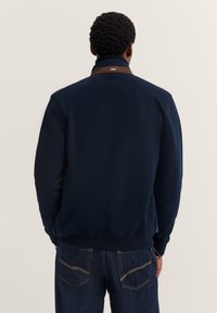 Uomo che indossa una giacca blu navy con un dettaglio del colletto marrone e jeans blu scuro, visto di spalle su uno sfondo chiaro e semplice.