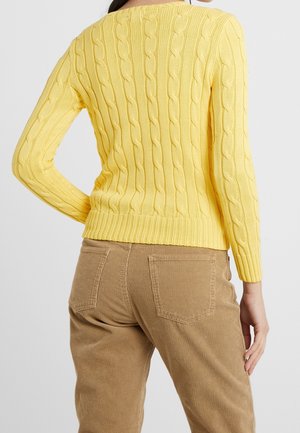 Femme portant un pull jaune en maille torsadée et un pantalon en velours côtelé marron, vue de dos avec une main posée sur la hanche.