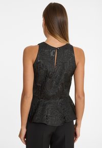 Femme aux longs cheveux bruns portant un haut peplum sans manches noir à motifs avec un détail de bouton en trou de serrure au dos, associé à un pantalon noir.