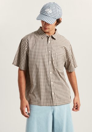 Chemise à manches courtes à carreaux marron et crème avec un devant boutonné et une poche poitrine, assortie à un short large bleu clair et une casquette à carreaux bleus.