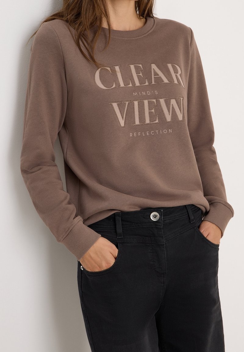 Femme portant un sweat-shirt marron à manches longues avec le texte « CLEAR VIEW » et un pantalon noir, les mains dans les poches, debout contre un mur clair.