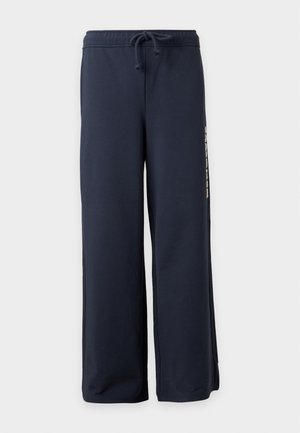 Hombre con chaqueta marrón claro, pañuelo verde a rayas en el cuello, pantalones azul marino, zapatos azules, sosteniendo un bolso de rayas rojas y azules en el hombro.