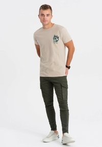 Beige tričko s krátkým rukávem s potiskem lebky a koruny na hrudi, v kombinaci s tmavě zelenými cargo kalhotami a bílými teniskami.