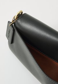 Pochette bracelet en cuir noir avec intérieur marron, anneau doré et étiquette noire attachée par une chaîne de perles dorées.