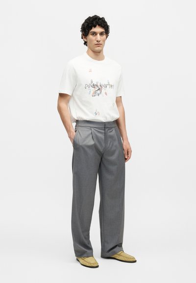 Άνδρας με κατσαρά μαλλιά που φοράει λευκό γραφικό μπλουζάκι, γκρι πιετέ παντελόνια, μπεζ μοκασίνια, στέκεται με το ένα χέρι στην τσέπη.
