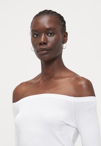 Hvit av-shoulder topp med lange ermer og et glatt, lett stoff. Modellen har kort flettet hår og enkle hoop øredobber.