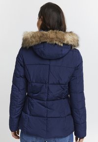 Marinblå pufferjacka med fuskpälsdetalj på huvan, med ett quiltat mönster och figurformad midja.