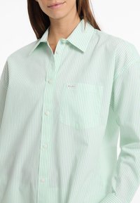 Lichtgroene en witte gestreepte blouse met een puntkraag, borstzak en lichte stof. Heeft witte knopen voor sluiting.