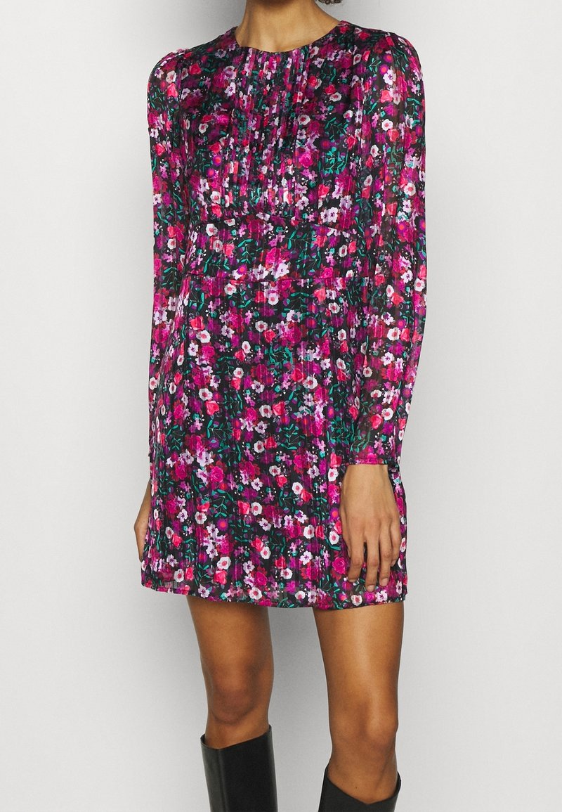 Femme portant une robe courte à manches longues à fleurs roses, violettes et blanches, associée à des bottes noires montantes jusqu'aux genoux, posant contre un mur blanc.