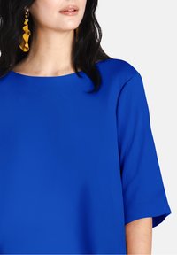Top blu royal con scollo rotondo e maniche corte, caratterizzato da una superficie liscia. Orecchini lunghi e ondulati di colore giallo creano un contrasto.