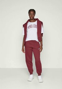 Sudadera marrón y joggers con un logo blanco, combinados con una camiseta blanca con letras rojas. El modelo está de pie contra un fondo claro.