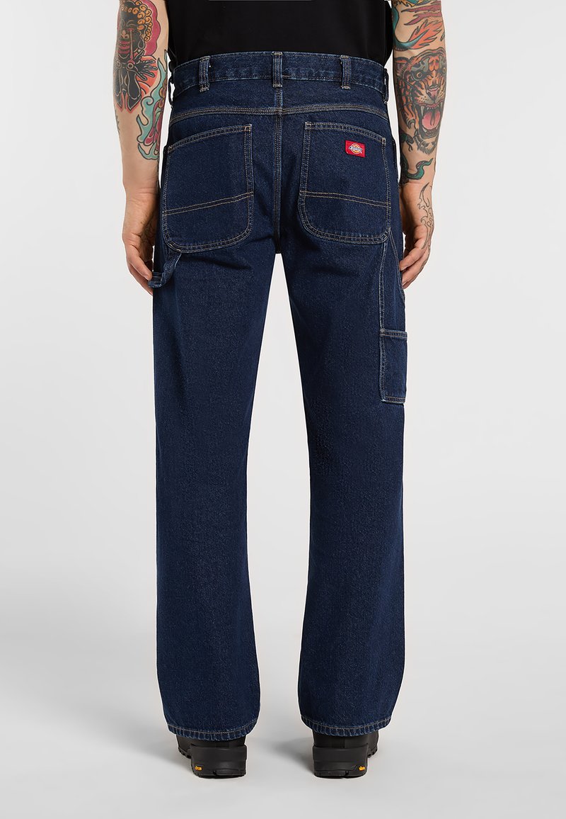 Set bagfra af en person iført mørkeblå denim tømrerbukser med store baglommer og sidelomme til værktøj, sorte sko og tatoveringer på begge arme.