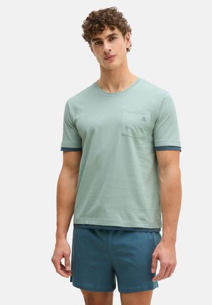 Junger Mann mit lockigem Haar, der ein hellgrünes T-Shirt mit kurzen Ärmeln und Brusttasche sowie dunkelgrüne Shorts trägt, vor einem schlichten Hintergrund stehend.