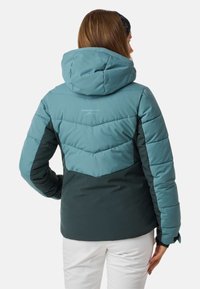Tealfarbene und dunkelgrüne Daunenjacke mit Chevron-Muster. Verfügt über eine Kapuze und eine gesteppte Textur, geeignet für kaltes Wetter. "Swedemount"-Logo sichtbar.