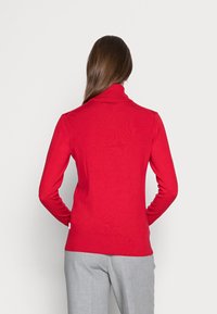 Rode coltrui van geribd stof, met lange mouwen en een aansluitende pasvorm. Draagt lichtgrijze broek.