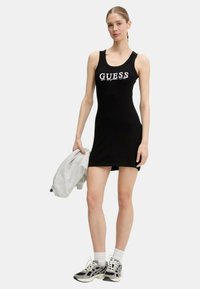 Abito a canotta nero a coste con logo "GUESS" bianco. Abbinato a una felpa grigia e sneakers nere/grigie, con calzini bianchi.