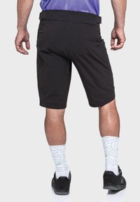 Schwarze Shorts mit glatter Textur, mittellanger Beinlänge bis zum Oberschenkel, seitlichen Taschen und einem engen Bund. Kombiniert mit gemusterten weißen Socken und schwarzen Schuhen.