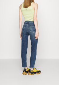 Mujer vestida con jeans de talle alto azules, top sin mangas de color verde claro y zapatillas gruesas amarillas y negras, de pie contra un fondo blanco liso.