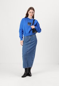 Felpa blu con un dettaglio tasca nera, abbinata a una lunga gonna di jeans e stivaletti neri alla caviglia. Design minimalista, vestibilità casual.