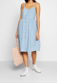 Ljusblå chambrayklänning med tunna axelband, knäppning framtill och insydd midja. Kombinerad med en rosa vävd tote-väska och vita sneakers.