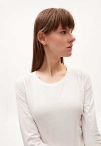 Weißes Langarmshirt mit rundem Ausschnitt, aus weichem Stoff gefertigt. Das Model zeigt ein Seitenprofil mit glattem, schulterlangem Haar.