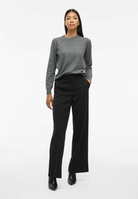 Chemise grise à manches longues associée à un pantalon large noir taille haute. Bottines à talons noires. Design simple et moderne.