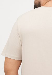 Licht beige T-shirt met korte mouwen, gladde textuur, ronde halslijn, met zichtbare stikdetails op de mouwen en schouders.
