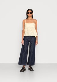 Top amarelo sem mangas com padrões brancos em gotas, combinado com jeans de corte largo em denim escuro e sandálias plataforma pretas. Design simples e casual.