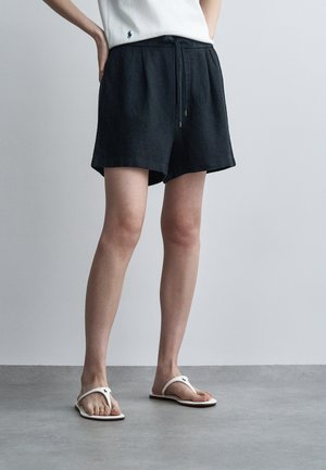 Άτομο που φοράει μαύρα σορτς με κορδόνι, λευκό αμάνικο τοπ και λευκά σανδάλια σιγαστήρες, στέκεται σε γκρι πάτωμα μπροστά σε απλό τοίχο.