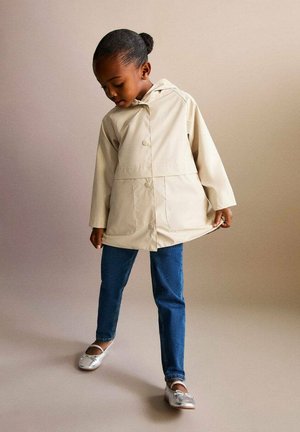 Lichtbeige waterdichte jas met een capuchon, drukknoopsluitingen en twee zakken, gecombineerd met blauwe jeans en zilveren ballerina's.
