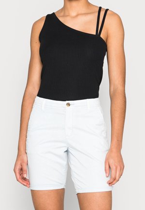 Femme portant un top noir côtelé sans manches à une épaule et un short blanc mi-cuisse avec bouton et passants pour ceinture.