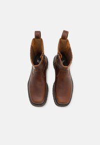 Botas de tobillo de cuero marrón con un acabado suave, que presentan un zipper en el lado y una suela negra robusta. Forro interior texturizado.